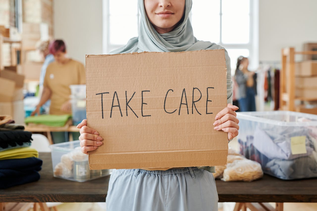 Smiling volunteer in hijab holding a Take Care sign at a charity event indoors.